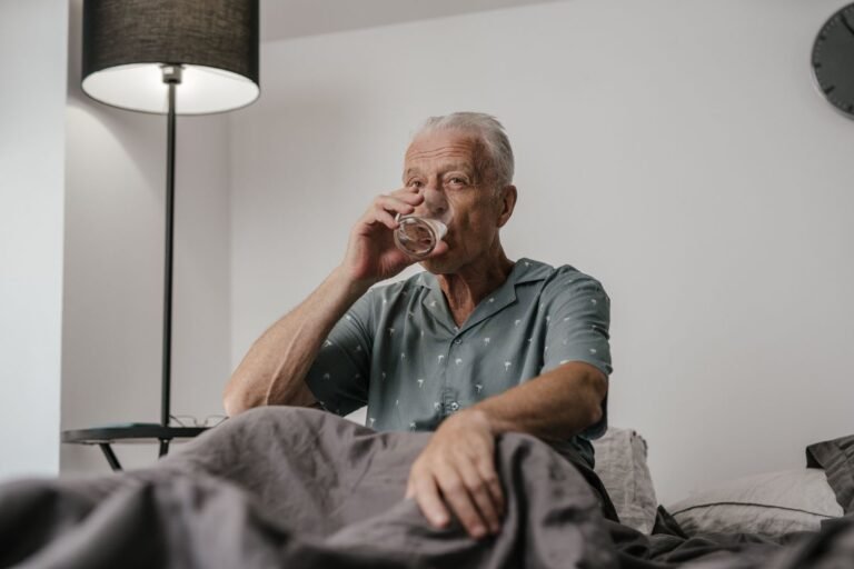 Un homme âgé boit un verre d'eau chez lui, illustrant la prévention de la déshydratation à domicile