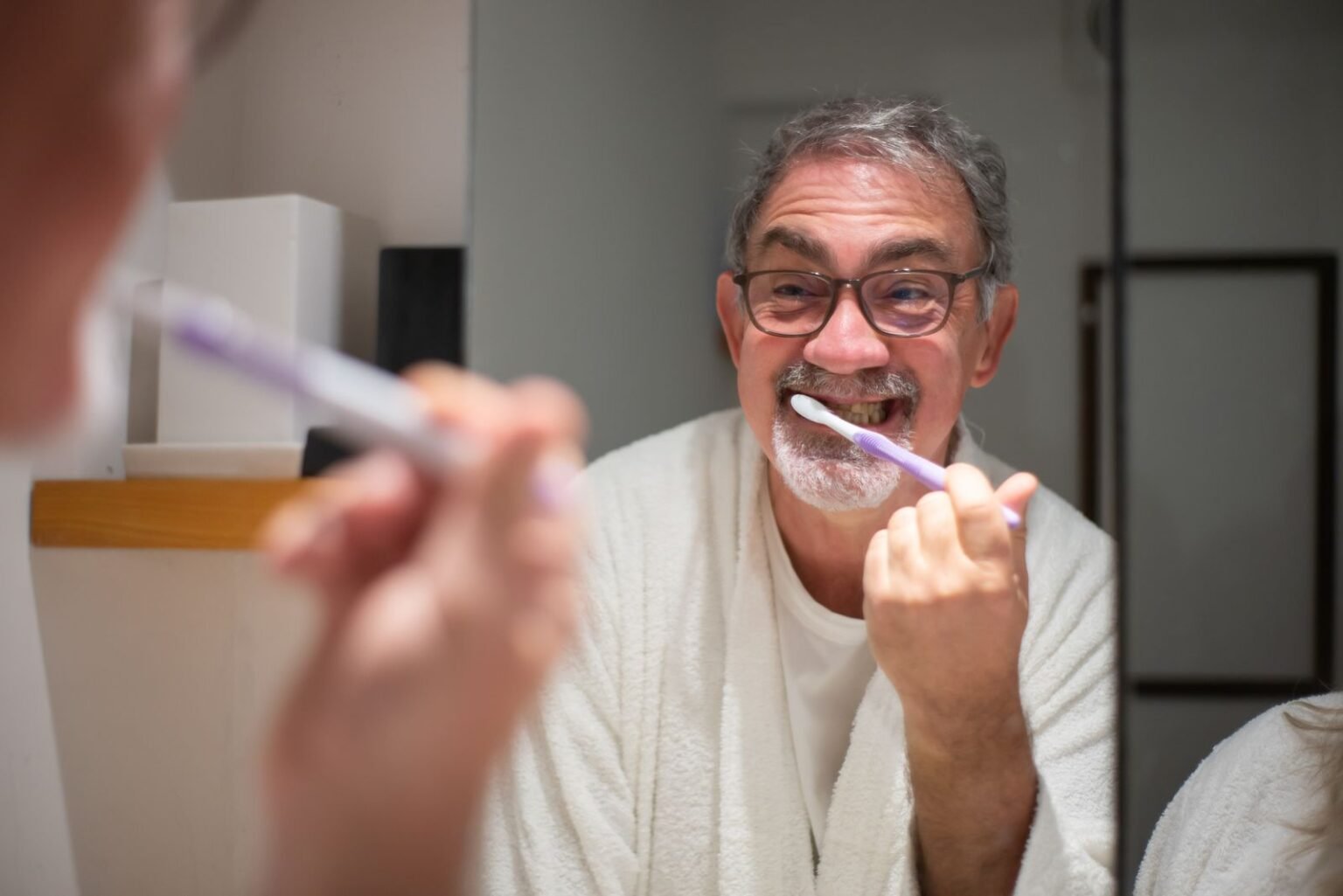 Homme âgé à lunettes se brossant les dents devant le miroir de sa salle de bain